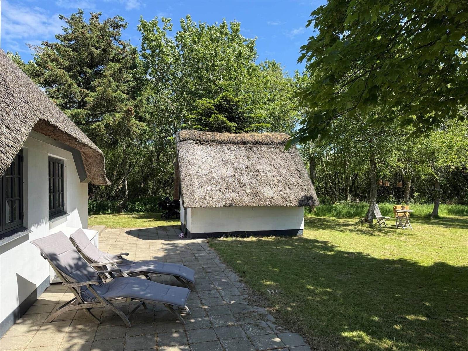 Zwei strohgedeckte Häuser mit Terrasse und Gartenbereich unter Bäumen.