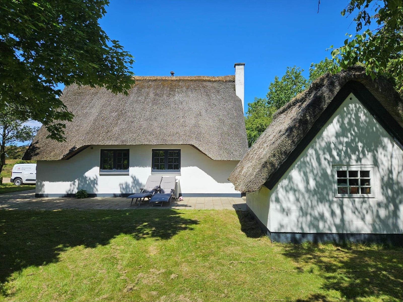 Zwei Häuser mit Strohdächern, Terrasse mit Liegestühlen und grüner Wiese unter blauem Himmel.
