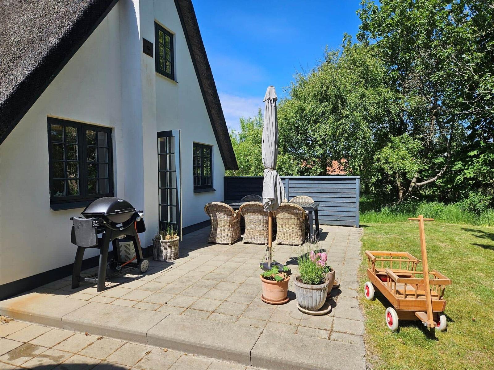 Terrasse mit Grill, Sitzgruppe und Kinderschubkarre vor einem Haus mit Strohdach.