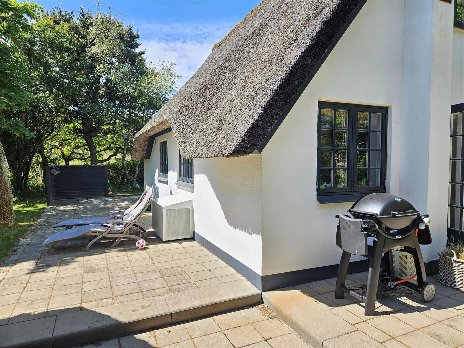 Haus mit Strohdach, Terrasse mit Liegestühlen und Grill.