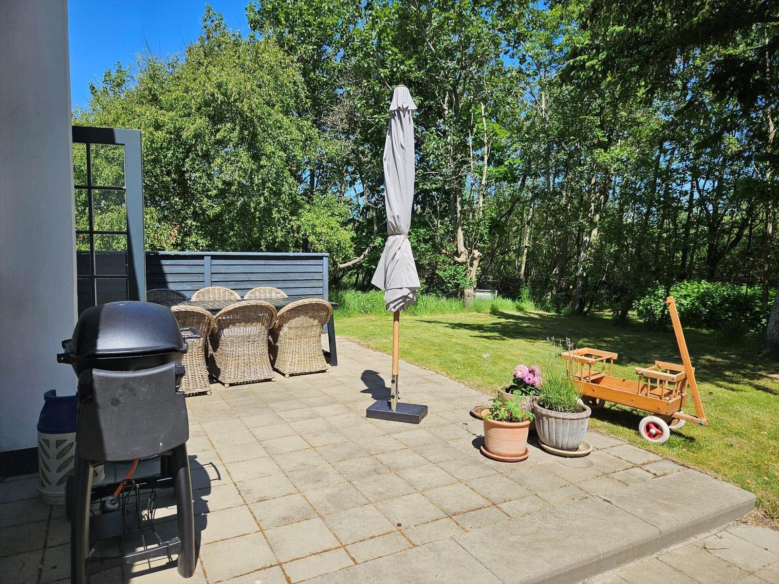 Terrasse mit Grill, Sitzgruppe, Sonnenschirm und Pflanzen. Hintergrund: grüner Garten mit Bäumen.