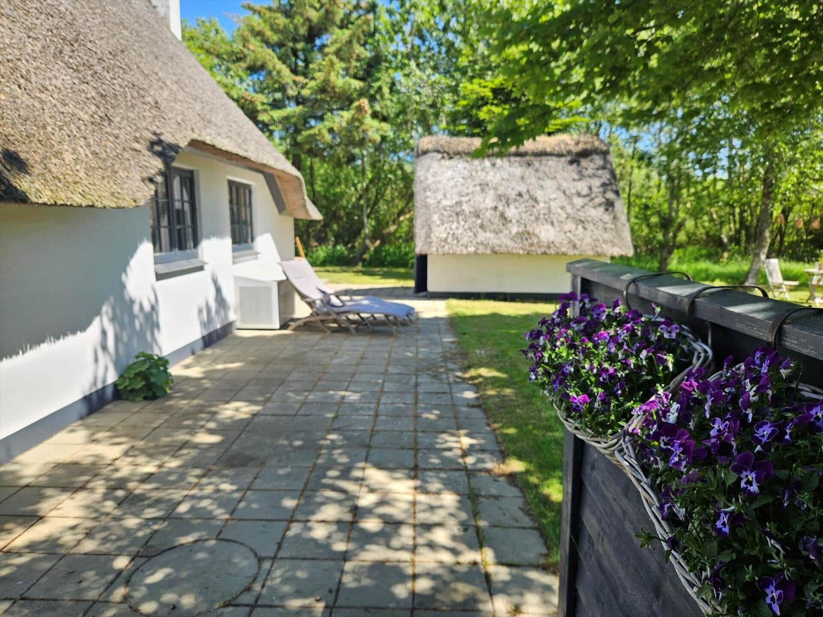 Hütte mit strohgedecktem Dach, Terrasse mit Liegestühlen und Blumenkästen an der Wand.