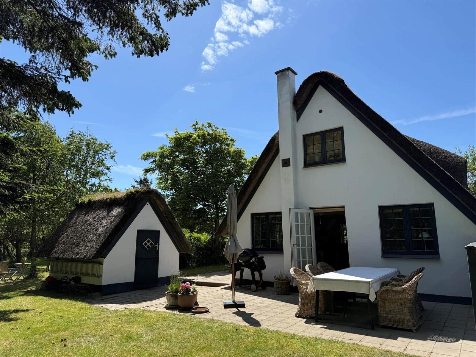 Haus mit Strohdach, Terrasse mit Tisch und Stühlen, Grill und Pflanzen im Garten.