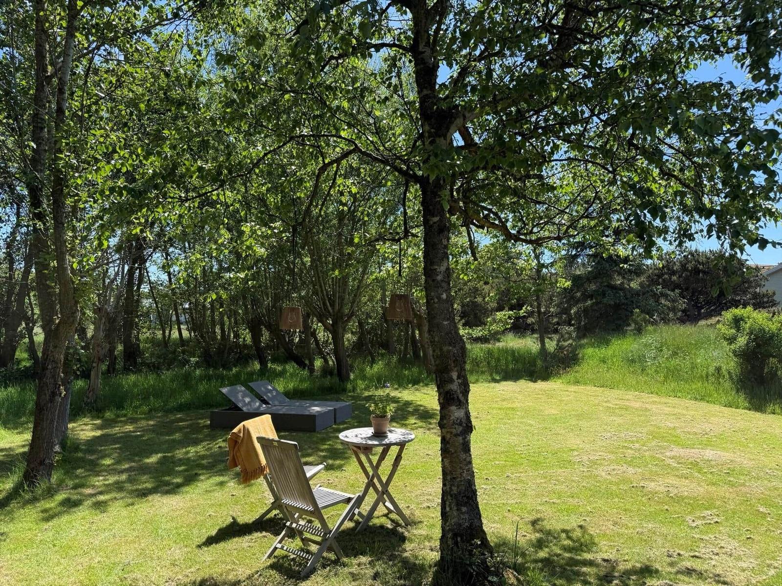 Garten mit Liege, Stuhl und Tisch unter Bäumen. Grasfläche und Wald im Hintergrund.
