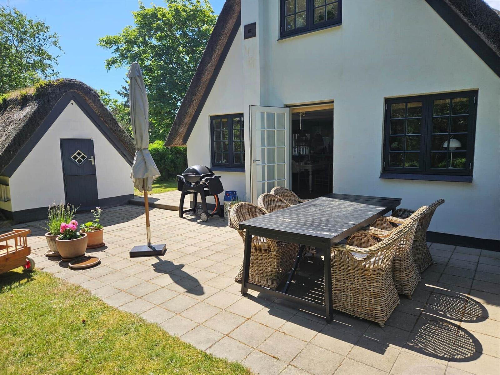 Terrasse mit Tisch, Stühlen, Grill und Sonnenschirm vor einem Haus mit Schieferdach.