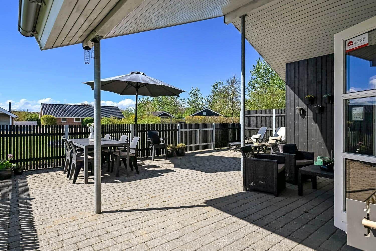 Terrasse mit Tisch, Stühlen und Sonnenschirm. Hinterhof mit Zaun und Bäumen.