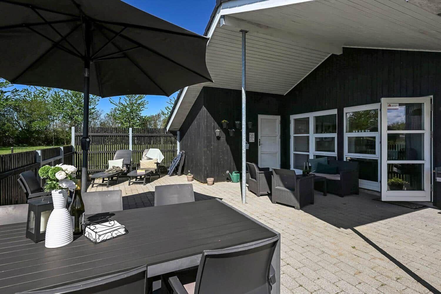 Terrasse mit Tisch, Stühlen und Sonnenschirm. Schwarze Hauswand und Glastüren sichtbar.