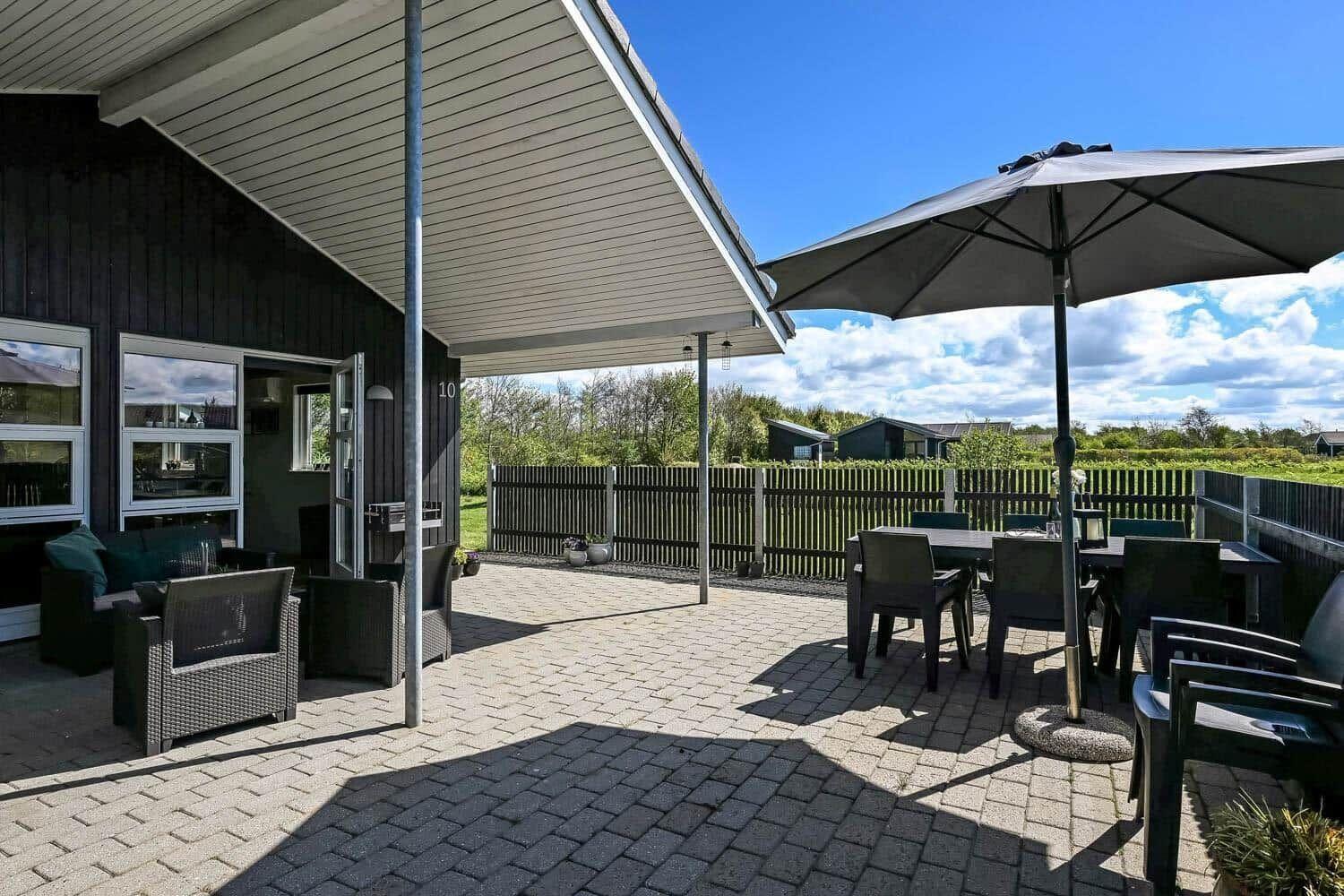 Terrasse mit Sitzgruppe, Tisch und Sonnenschirm. Hintergrund: Zaun und Grünflächen.