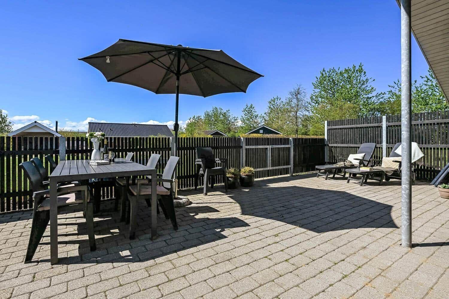 Terrasse mit Tisch, Stühlen und Sonnenschirm. Hintergrund: Holzzaun und Bäume.