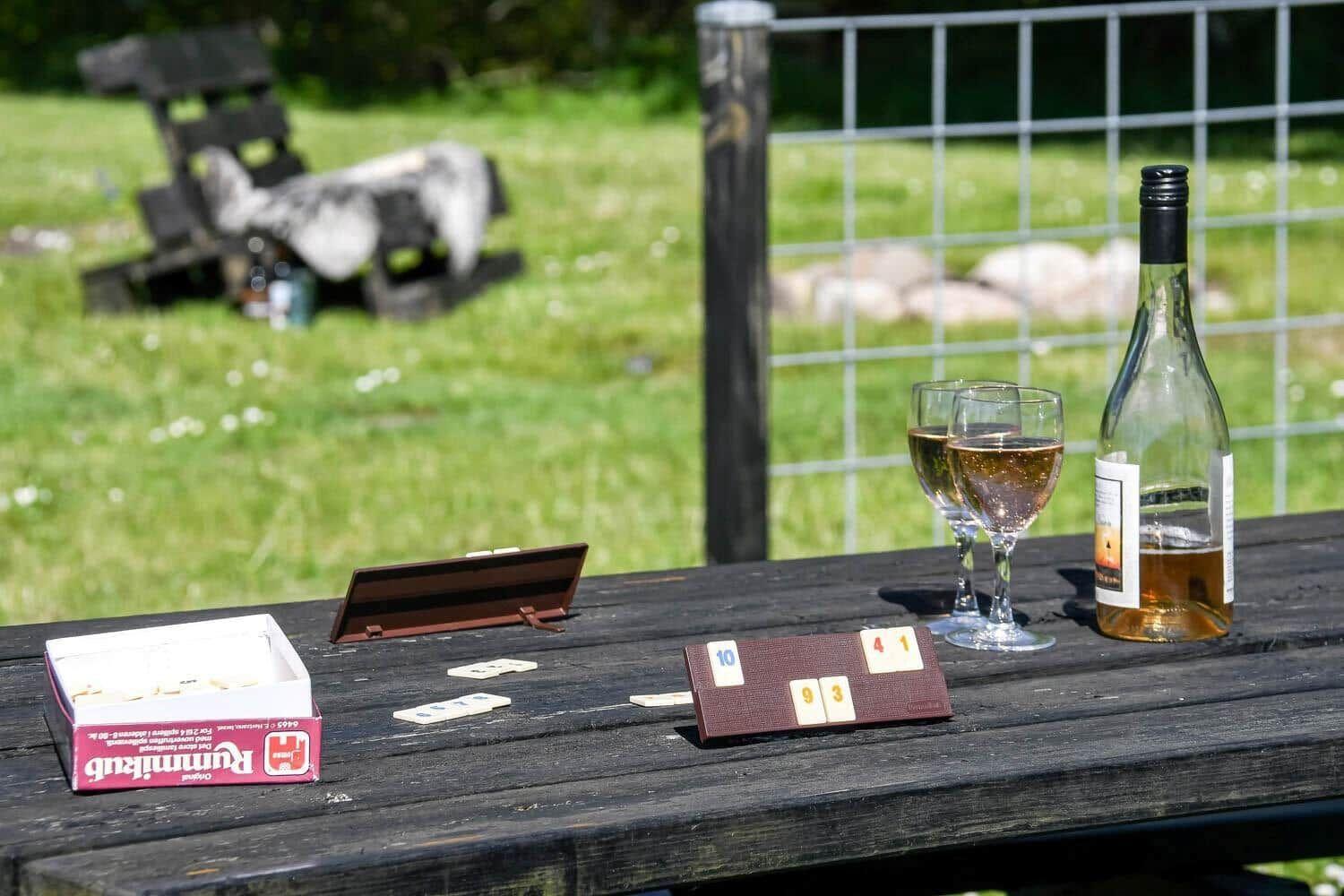 Holztisch mit Rummikub-Spiel, zwei Gläser und einer Weinflasche im Freien.