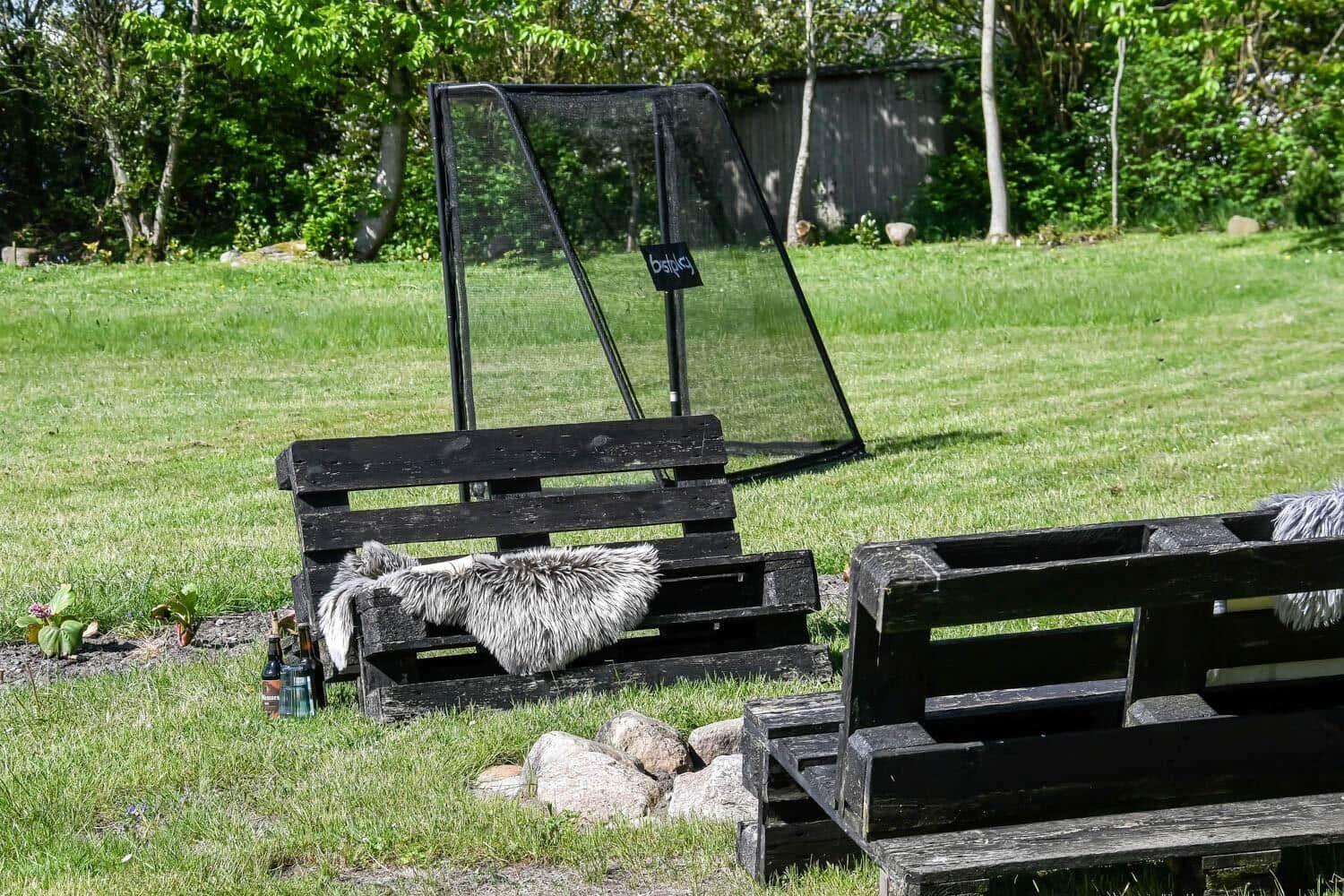 Zwei Palettenbänke mit Felldecken stehen auf einem Rasen. Hinter ihnen steht ein Schlagballnetz.