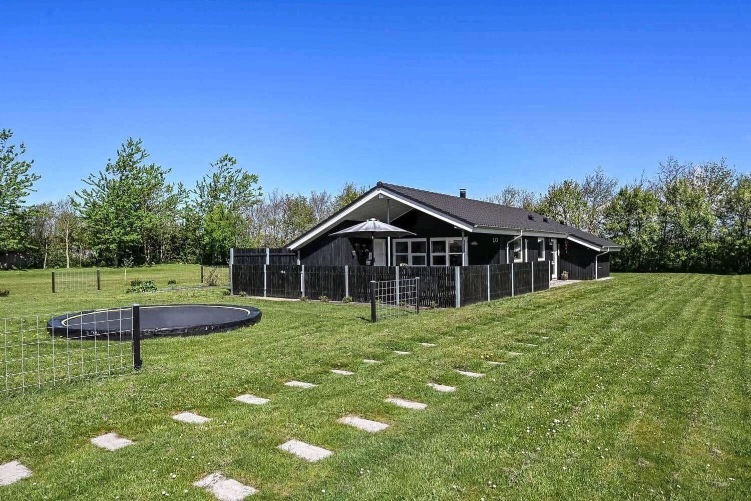 Schwarzes Haus mit Terrasse, Trampolin und Rasenfläche. Hinterhof mit Zaun und Bäumen.