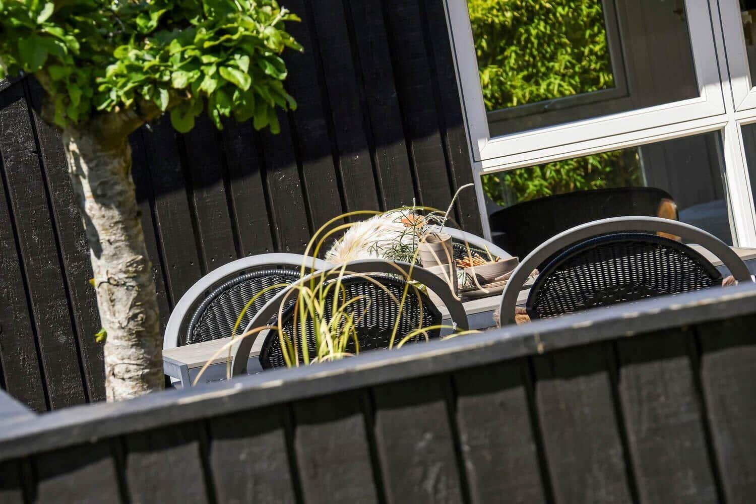 Terrasse mit Tisch, Stühlen und Pflanzen vor dunklem Holzzaun und Fenster.