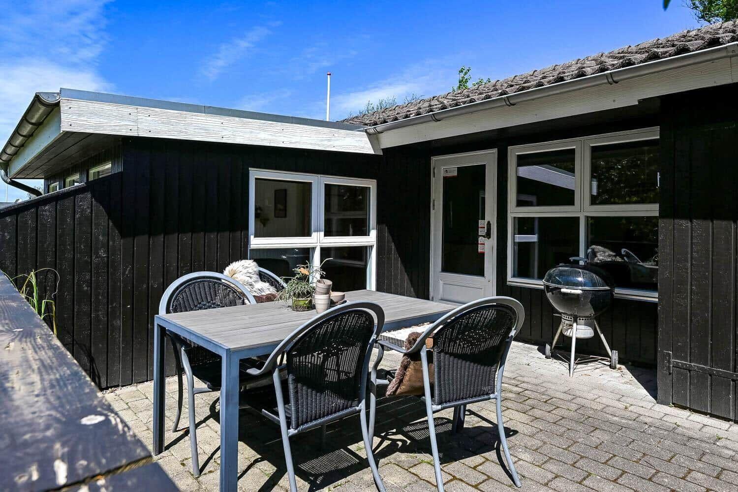 Terrasse mit Tisch, Stühlen und Grill vor einem Haus mit schwarzer Fassade.