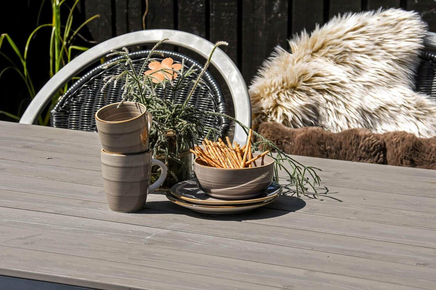 Holztisch mit Tassen, Schale und Pflanze im Garten. Hintergrund: Stuhl mit Felldecke.
