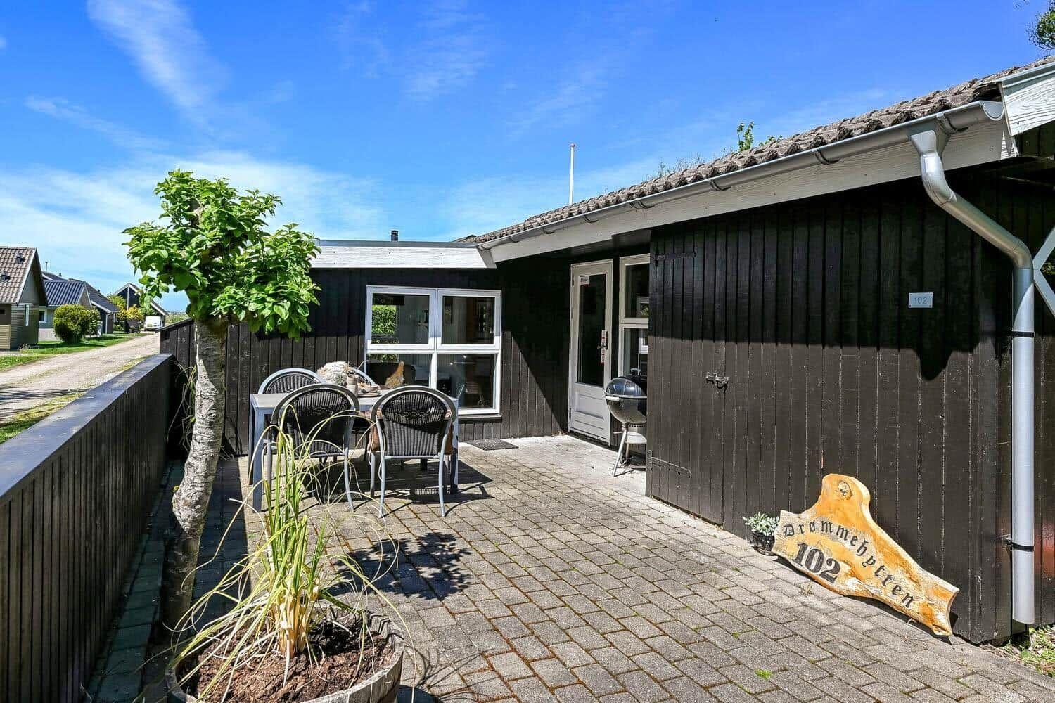 Haus mit Terrasse, Tisch, Stühlen und Grill. Holzschild mit Hausnummer 102.
