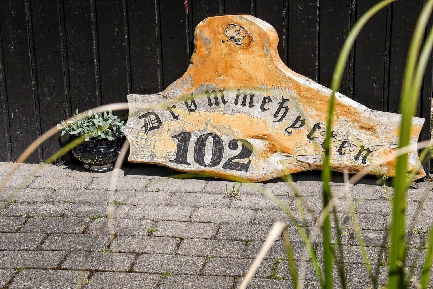 Holzschild mit der Aufschrift Drommehetten 102 vor einer schwarzen Holzwand.