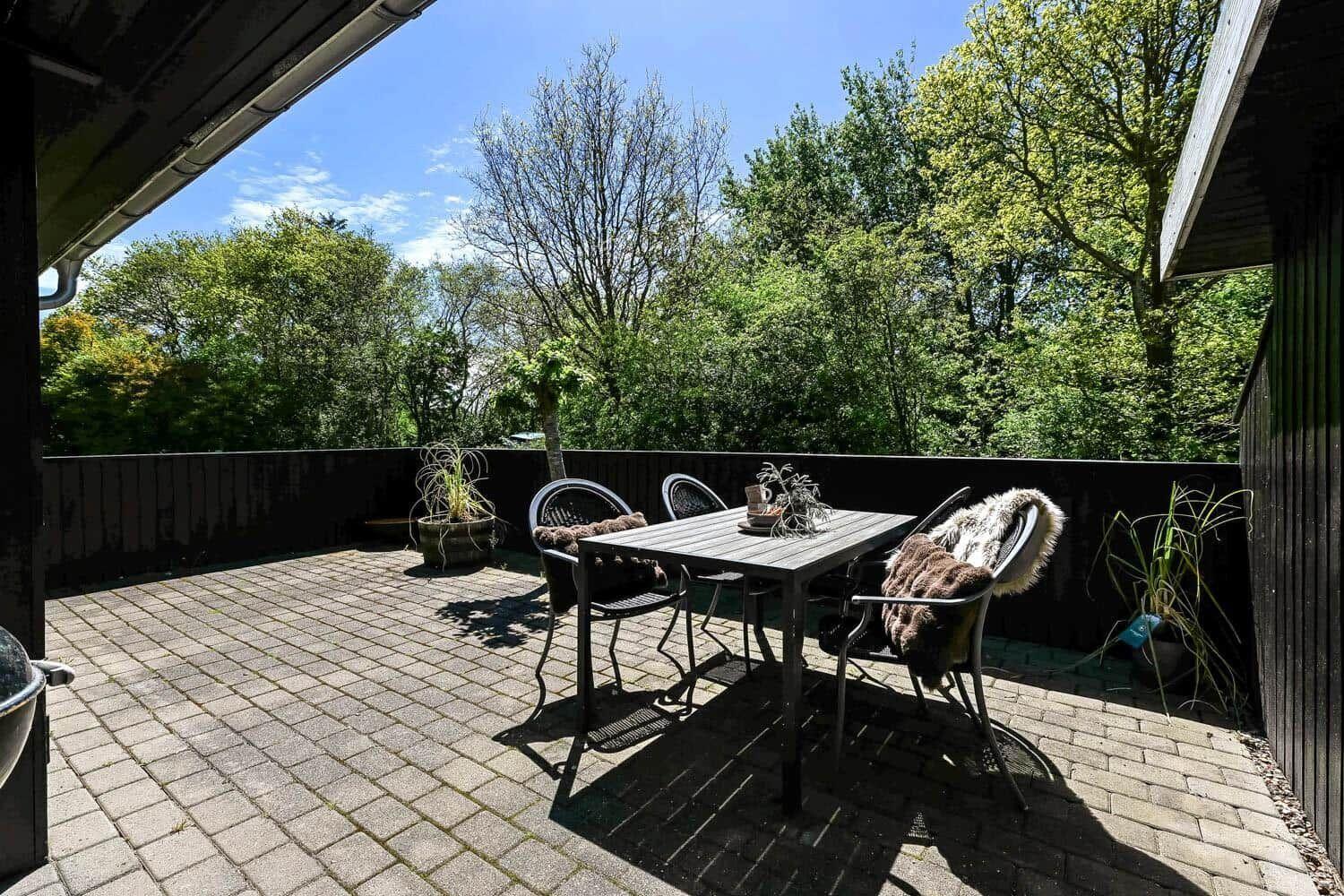 Terrasse mit Tisch, Stühlen und Pflanzen. Blick auf grüne Bäume und einen klaren Himmel.