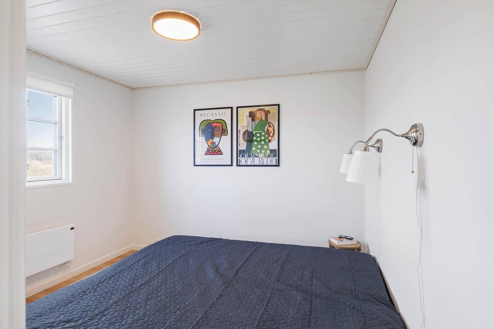 Schlafzimmer mit Bett, Wandbildern und Fenster. Deckenleuchte und Stehlampe.