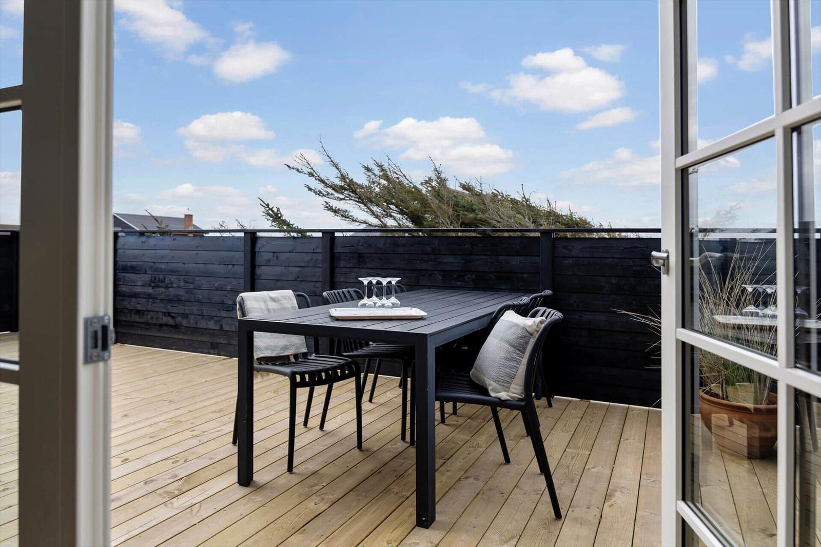 Terrasse mit Tisch und Stühlen, Holzdecke und schwarzer Holzabtrennung. Blick auf Bäume und Himmel.