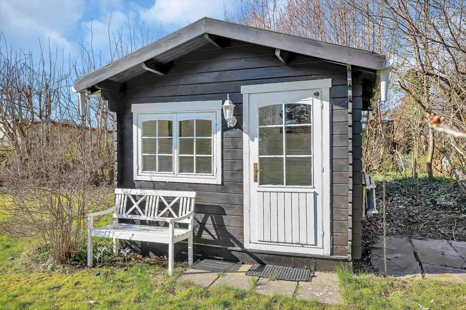 Ein kleines Haus mit weißer Tür und Fenster, einem weißen Gartenbank und einem grauen Dach.