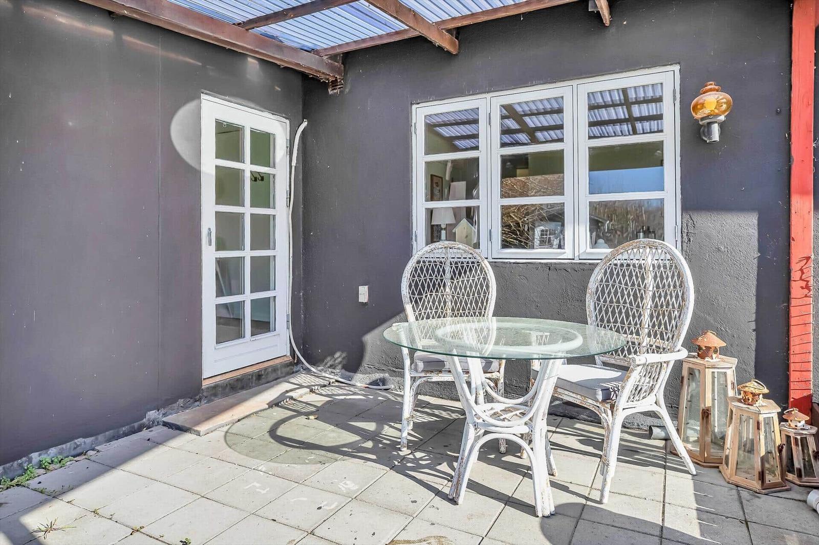 Terrasse mit Tisch und zwei Stühlen, graue Wand, Fenster und Tür.