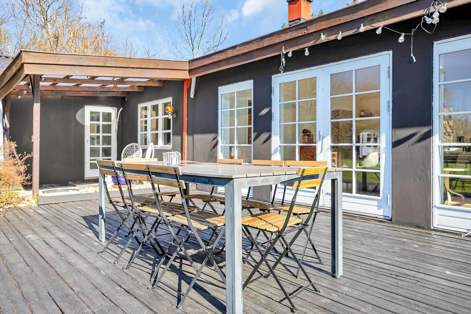 Terrasse mit Holztisch und Stühlen vor einem Haus mit weißen Fenstern.