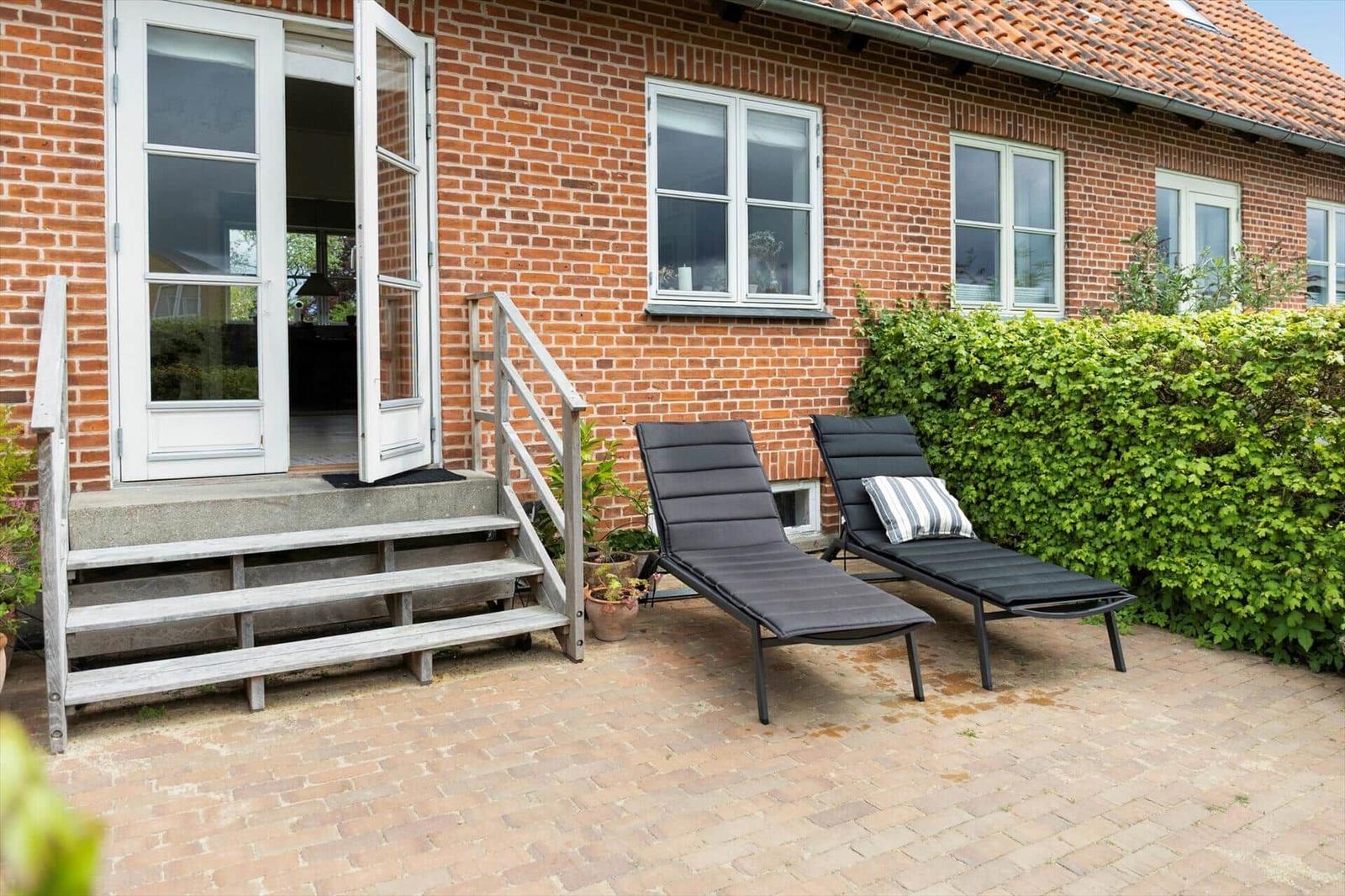 Red brick house with terrace, two lounge chairs, and green hedge.