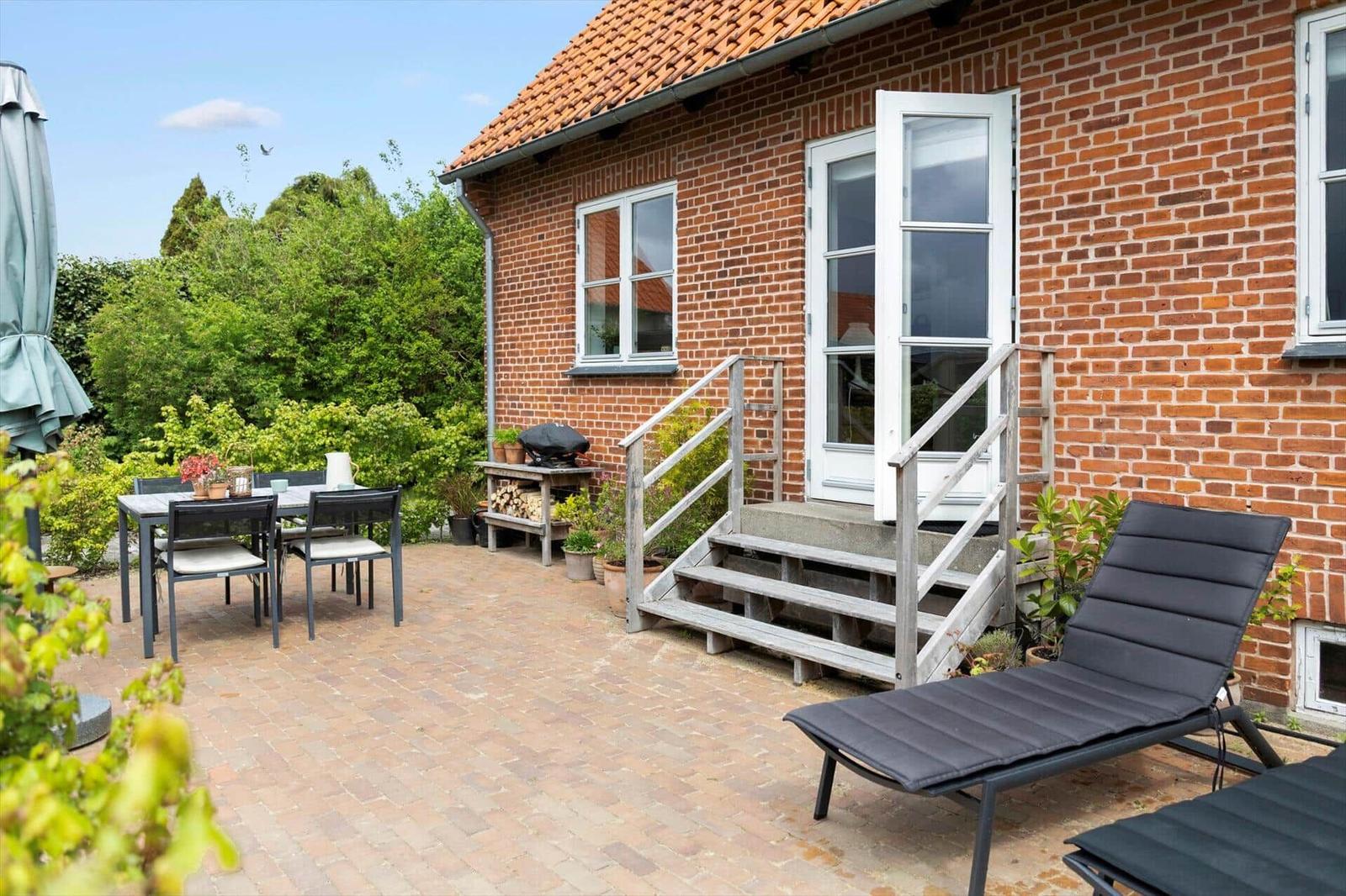 Terrace with table, chairs, lounge chairs, and grill in front of a brick house.