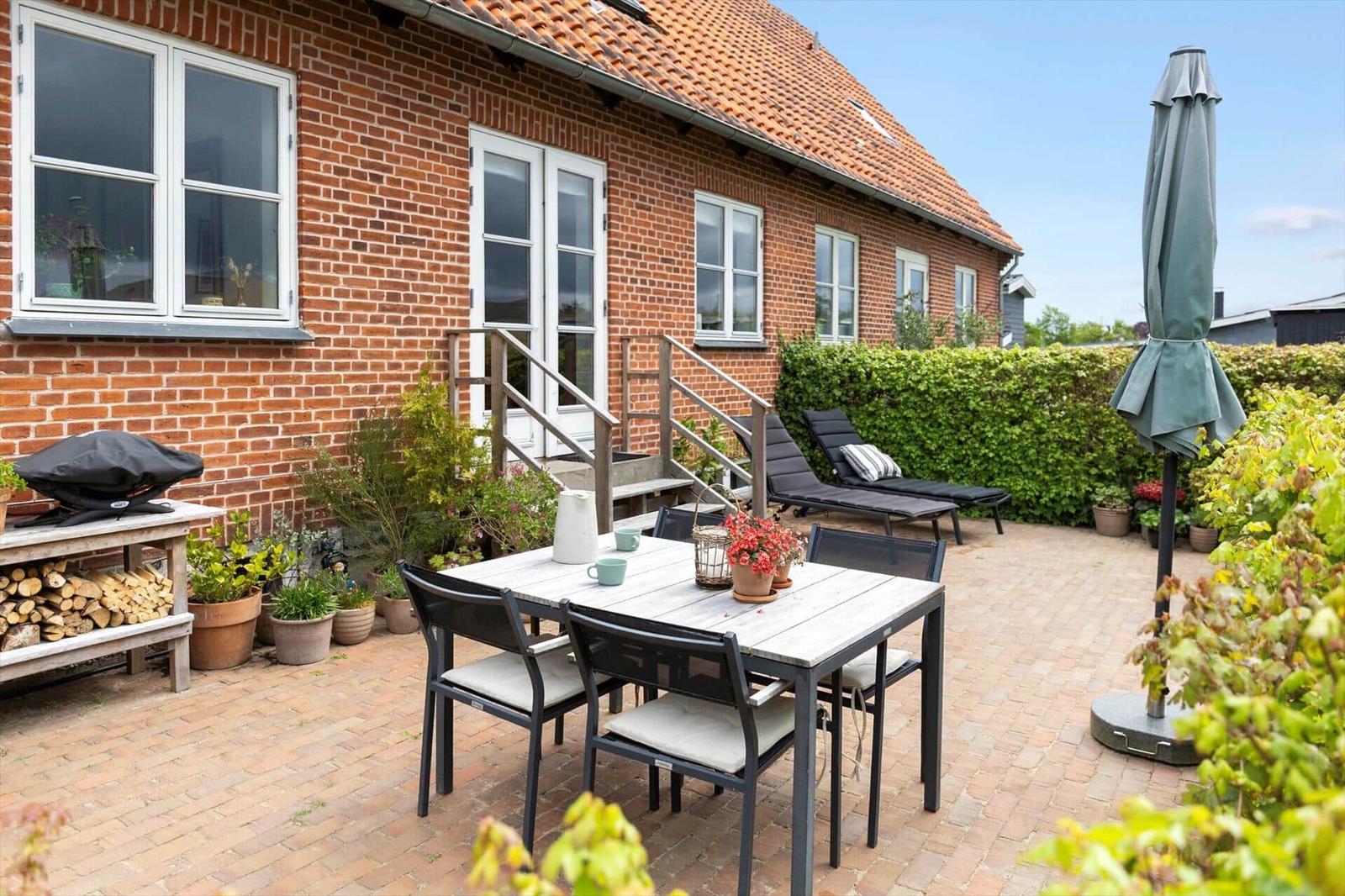 Backyard with table, chairs, sun loungers, and grill. Red brick building with white windows.
