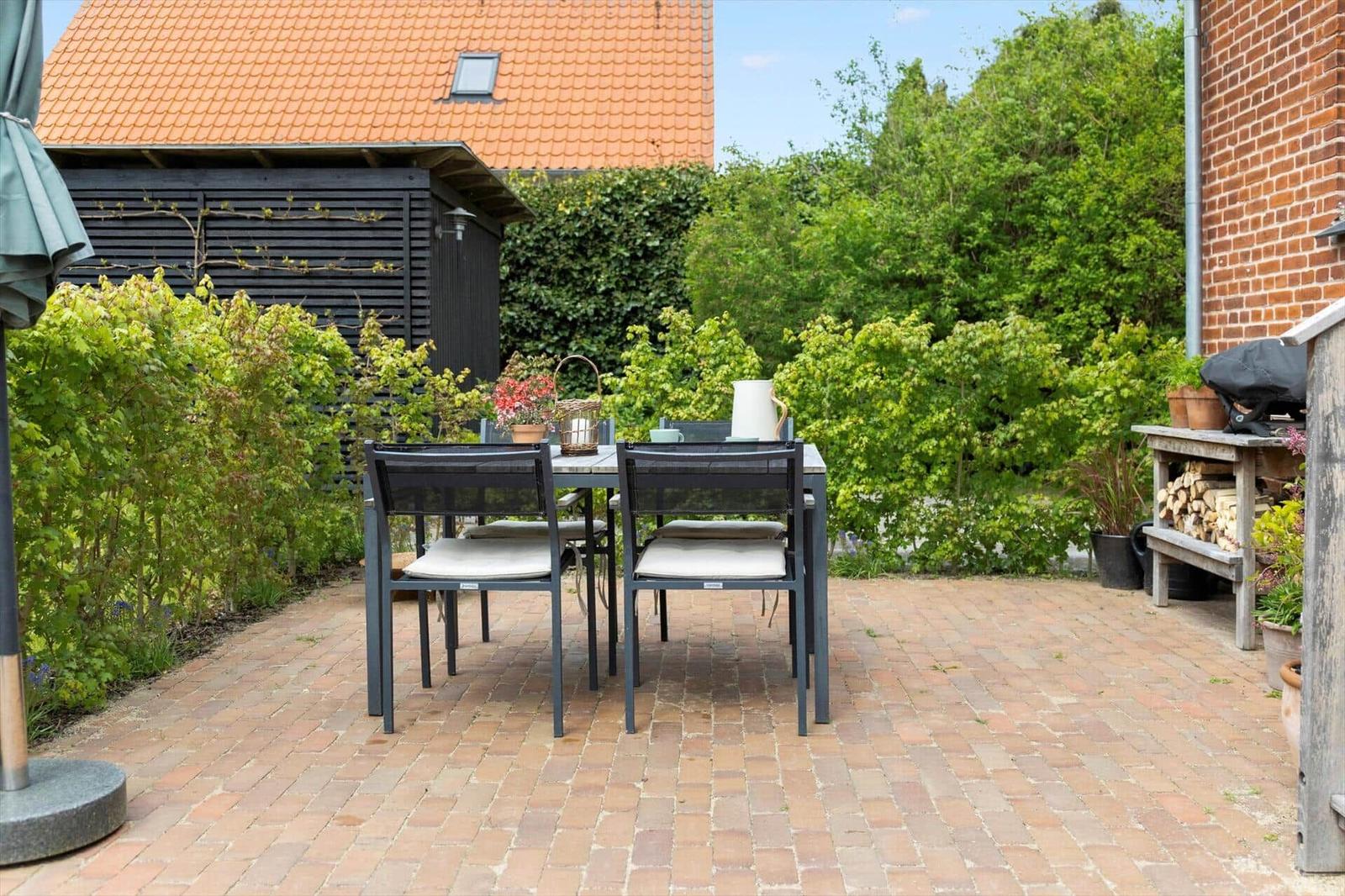 Terrace with table and chairs, surrounded by greenery and a grill.