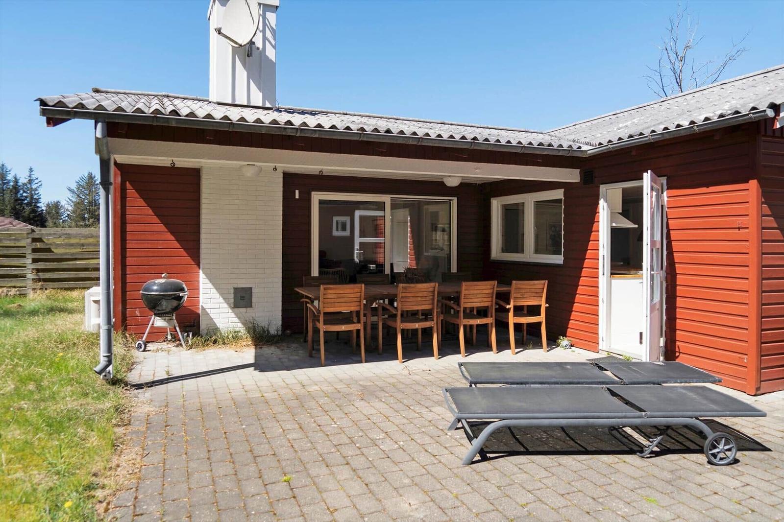 Roter Holzhaus mit Terrasse, Grill und Liegen. Durchgangstür und Fenster sichtbar.