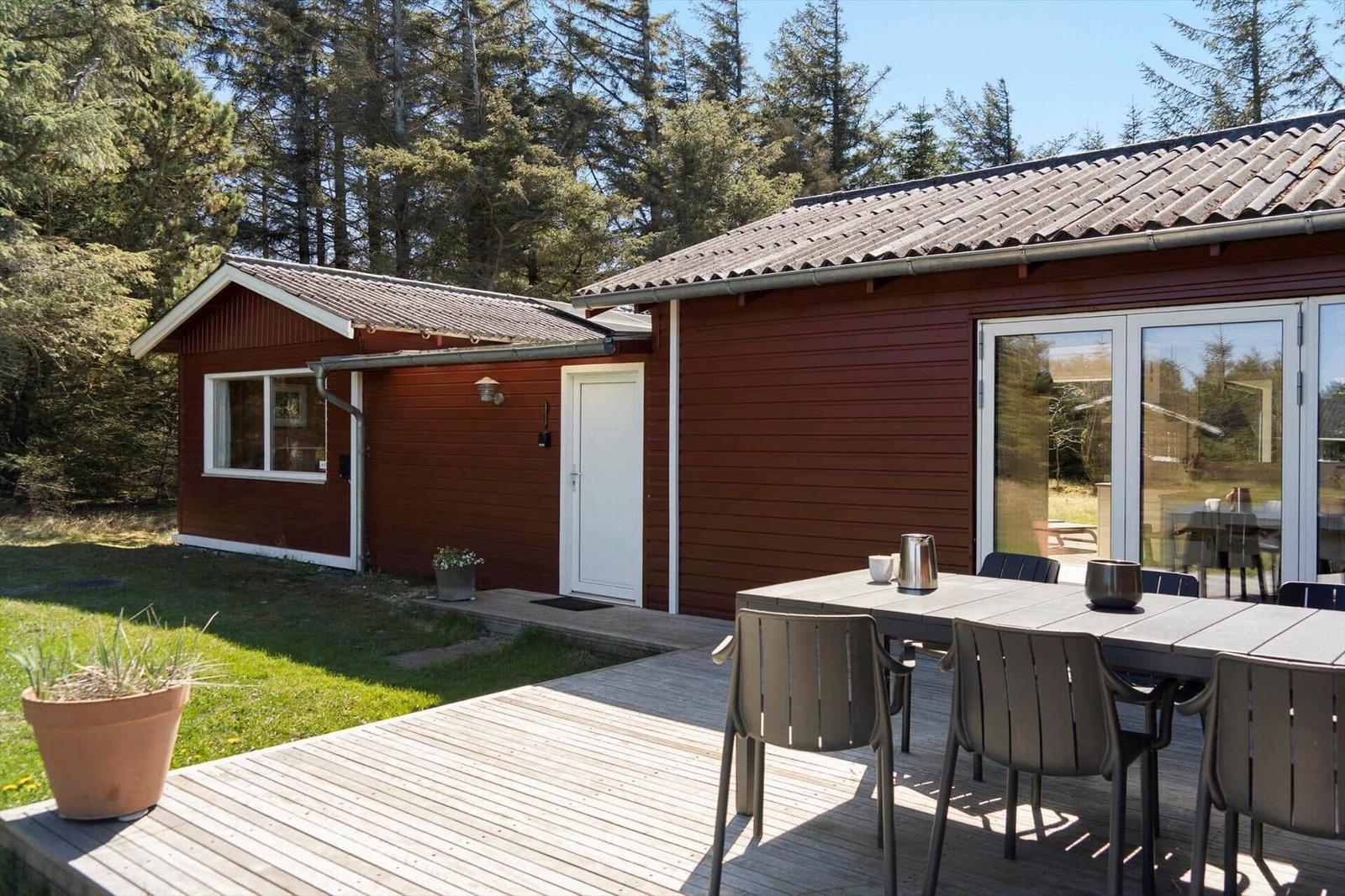 Rotes Ferienhaus mit Terrasse, Tisch und Stühlen. Hintergrund: Wald.