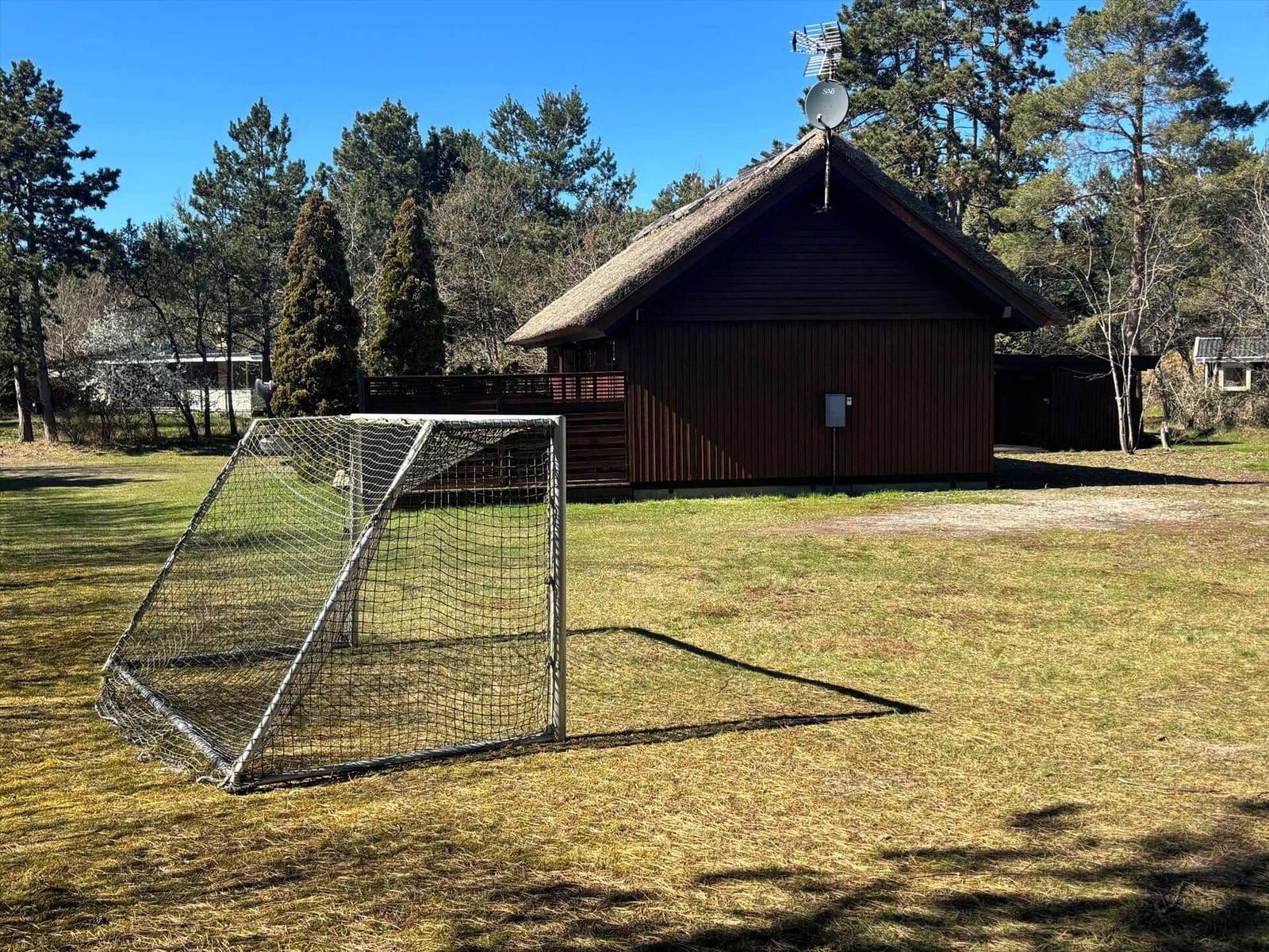 Holzhütte mit Dach aus Stroh, Fußballtor im Vordergrund, umgeben von Bäumen und Gras.