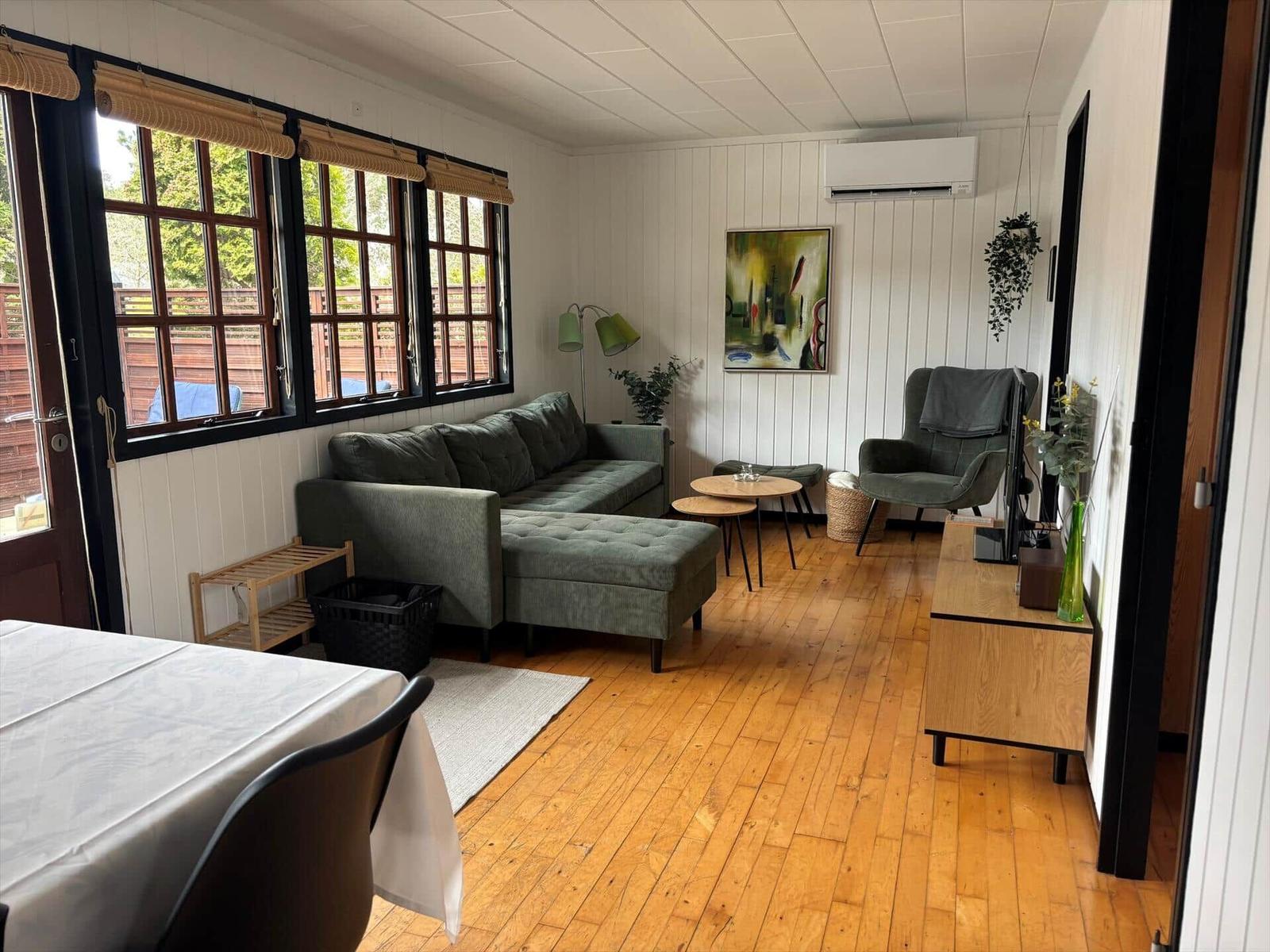 Wohnzimmer mit Sofa, Sessel, Tisch und Holzboden. Fenster mit Jalousien. Wandbild und Pflanzen.