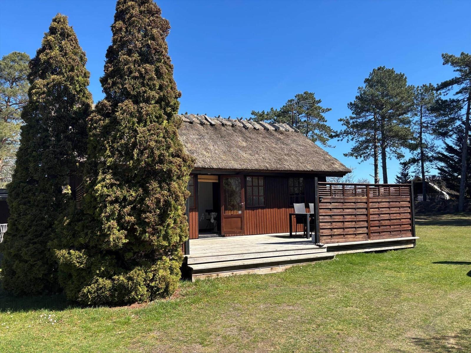 Holzhütte mit Strohdach und Terrasse. Umgeben von Bäumen und Gras.