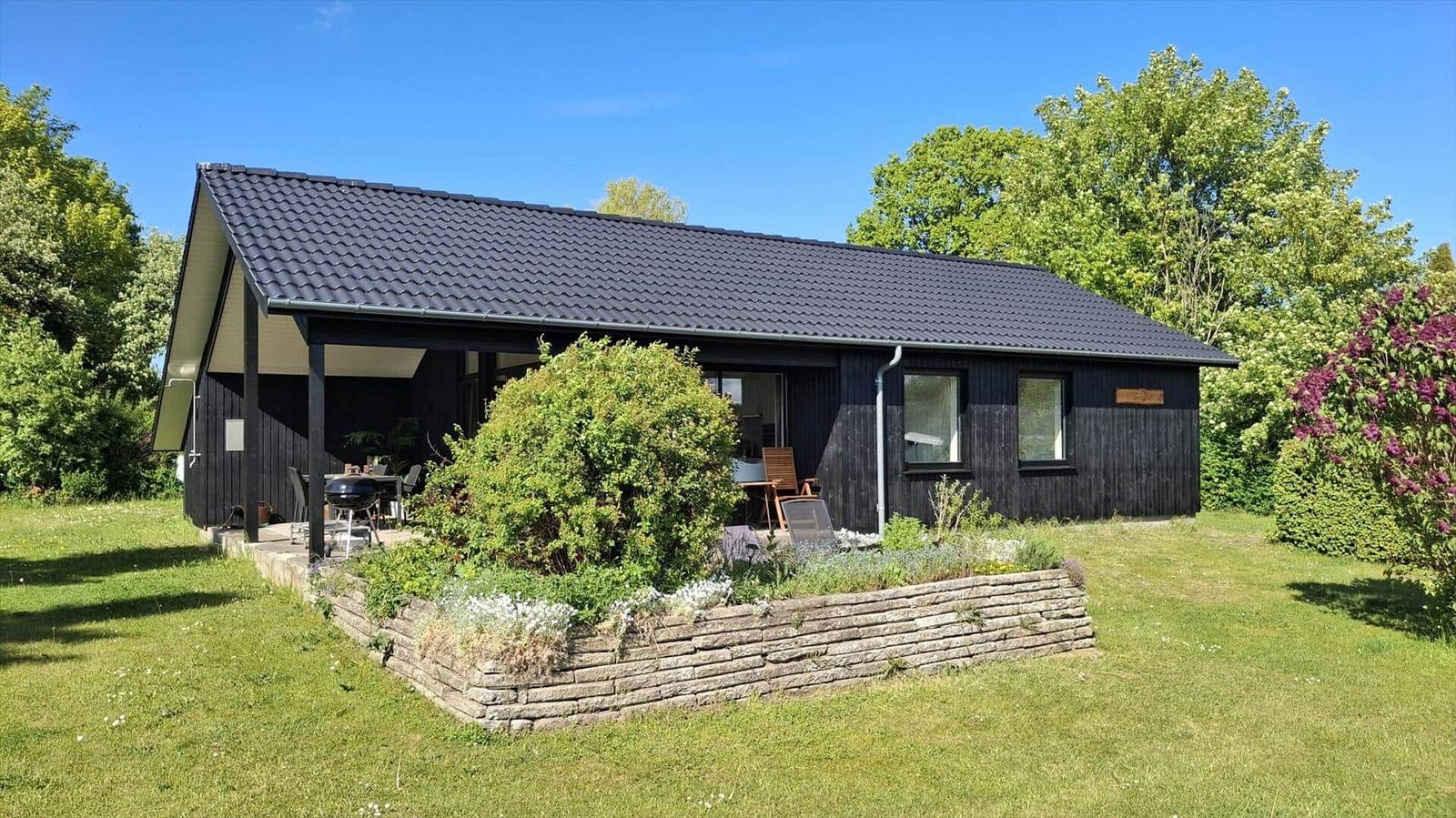 Black wooden house with terrace, grill, and garden. Surrounded by trees and shrubs.