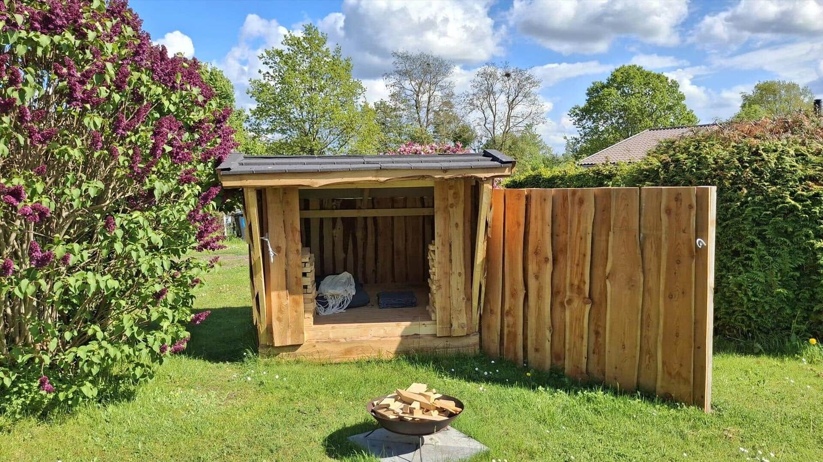 Wooden structure with open roof and wooden wall. Inside, wood piles and a cushion are visible.