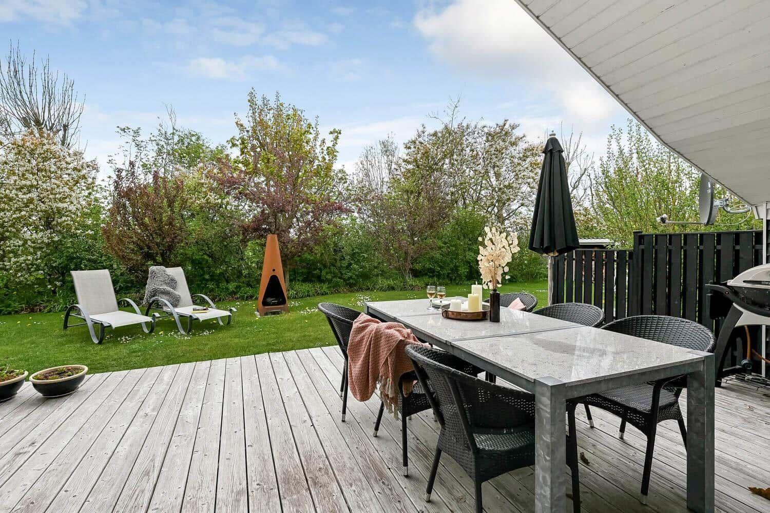 Terrasse mit Tisch, Stühlen, Liegestühlen und Grill. Hintergrund: grüner Garten mit Bäumen.