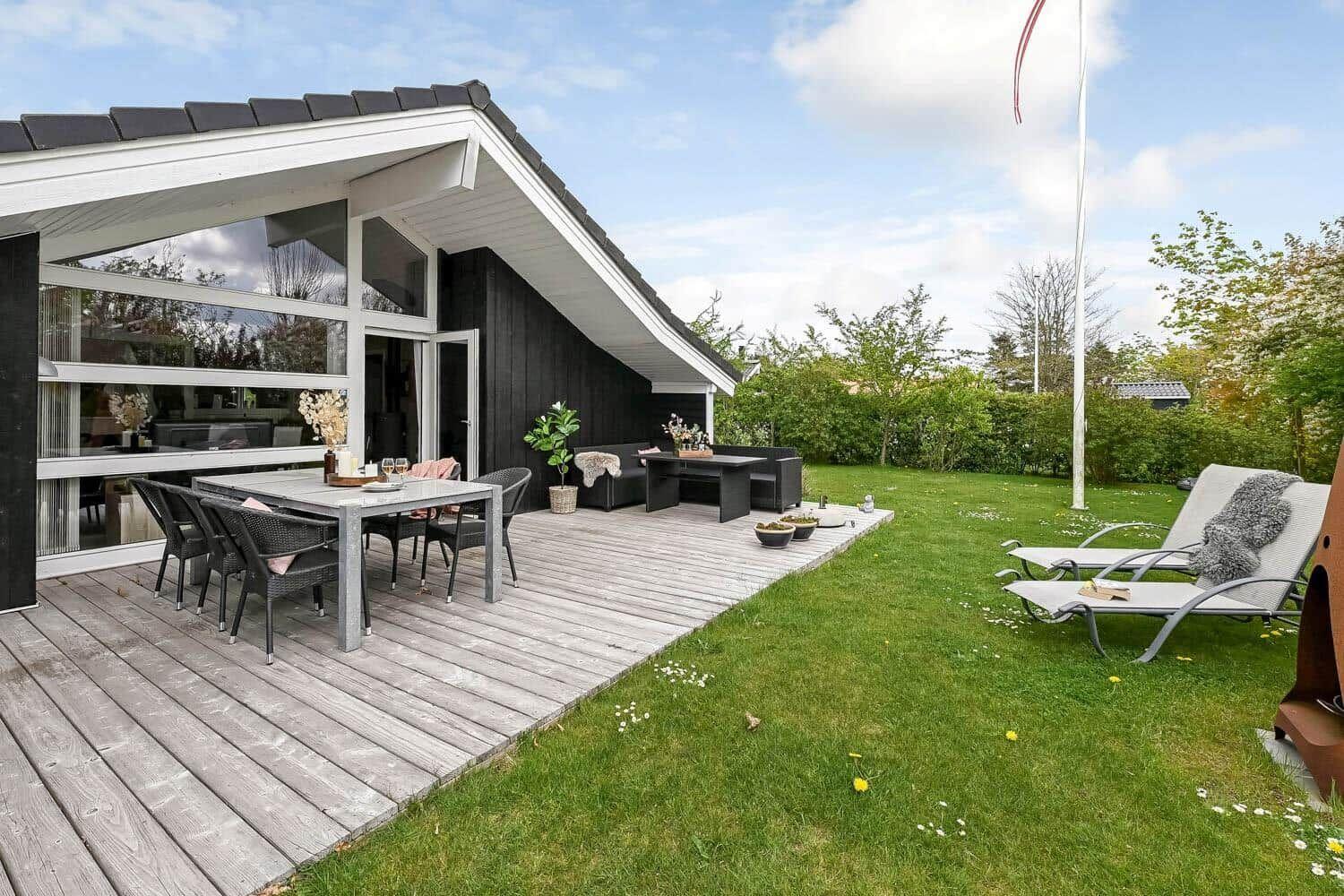 Haus mit großem Holzterrasse, Gartenmöbeln und Liegestühlen im Gras.