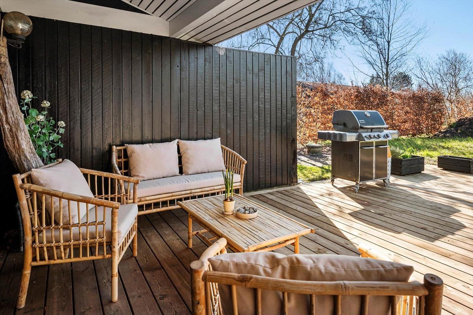 Terrasse mit Bambusmöbeln, Grill und Holzdecke. Hintergrund: Hecke und Bäume.