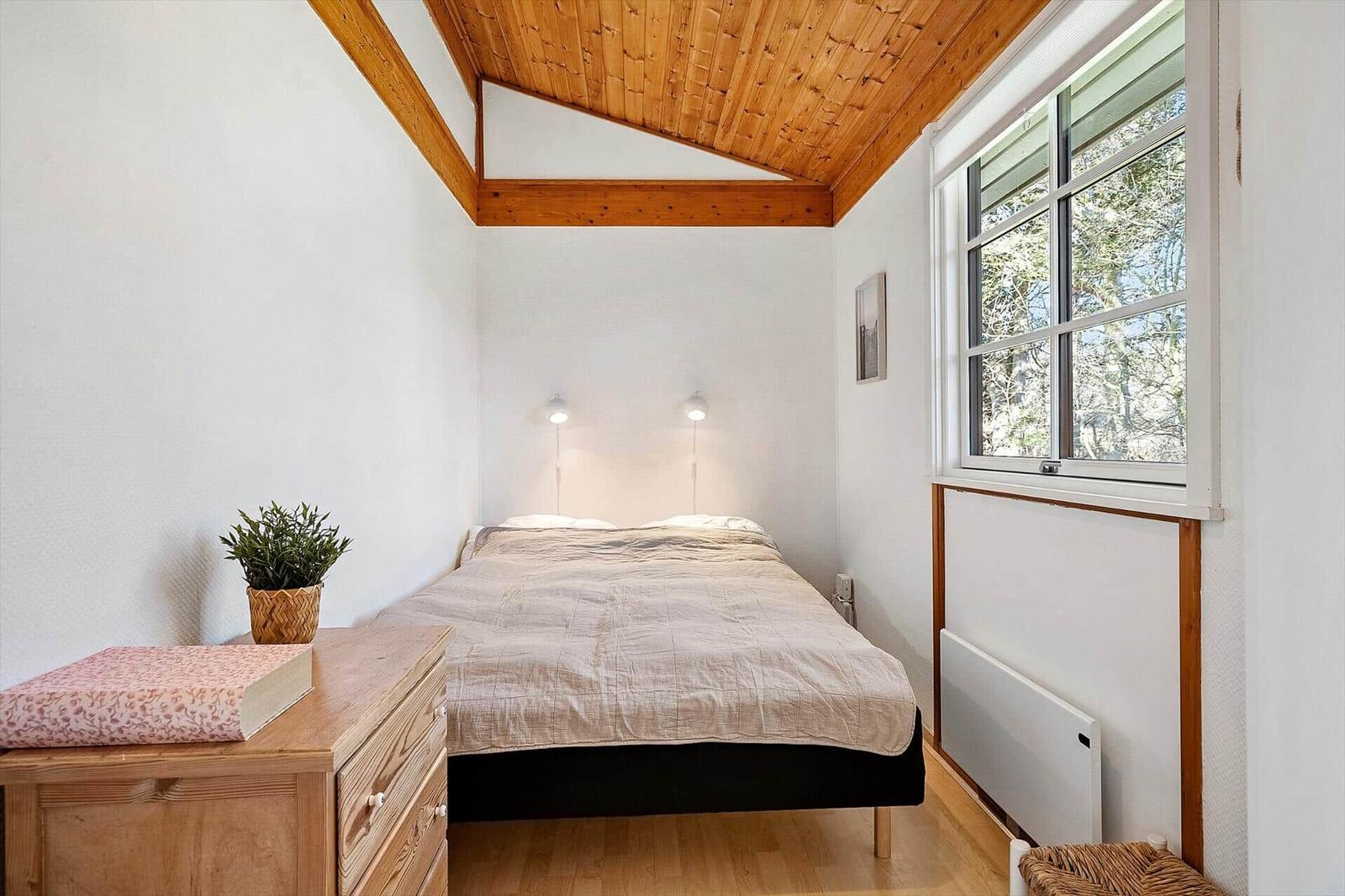Schlafzimmer mit Bett, Nachttisch, Pflanze und Fenster mit Blick auf Bäume.