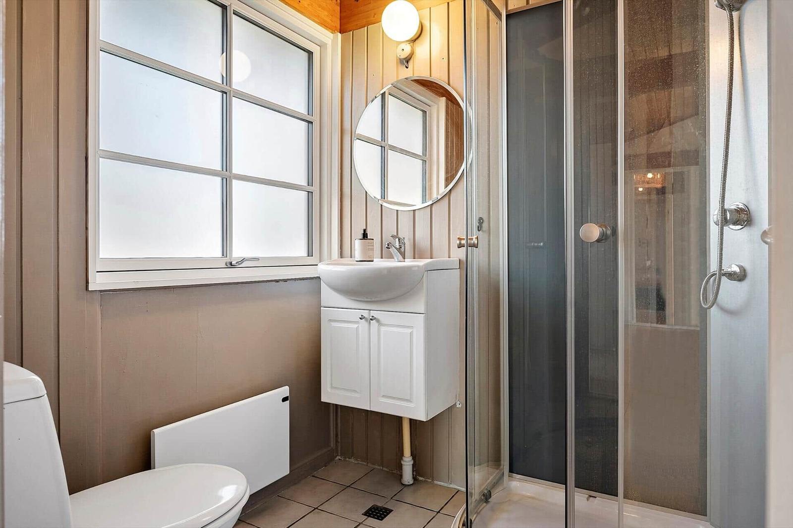 Badezimmer mit Dusche, Waschbecken und Toilette. Holzwände und Fenster sichtbar.