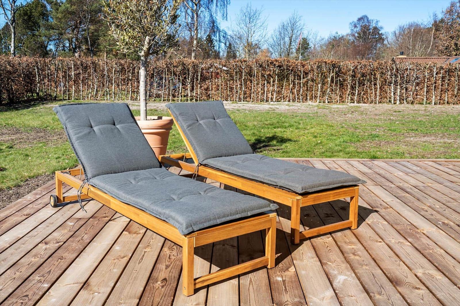 Zwei Liegestühle mit Kissen stehen auf einer Holzterrasse. Hintergrund: Gras, Heckenzaun und Bäume.