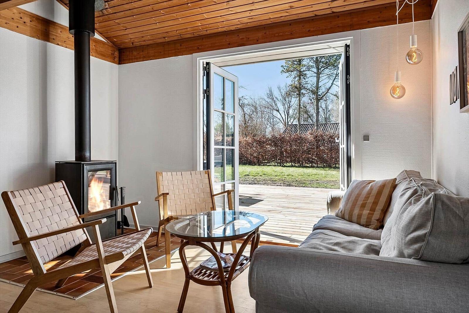 Wohnraum mit Kamin, Sofa und Tisch. Tür führt auf Terrasse mit Blick auf Garten.