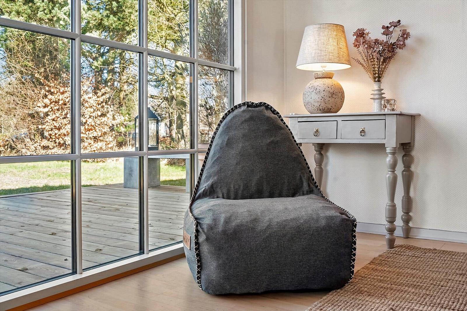 Ein grauer Sitzsack steht vor einem großen Fenster. Neben ihm steht ein Tisch mit Lampe und Vasen.