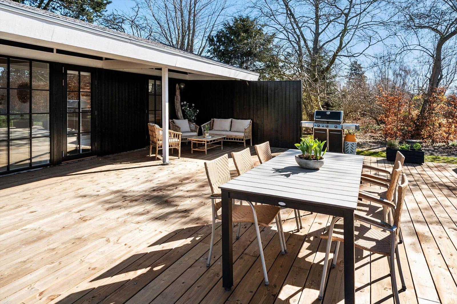 Terrasse mit Esstisch, Grill und Sitzbereich unter Pavillon. Holzdecke und Bäume im Hintergrund.