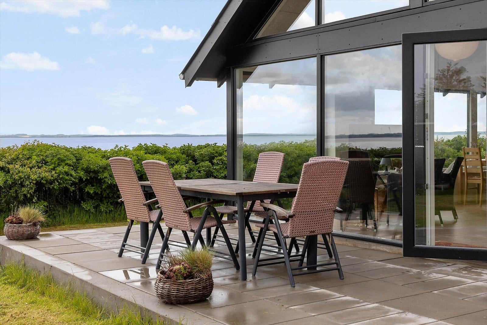 Terrace with table and chairs. View of water and greenery. Glass front to interior.
