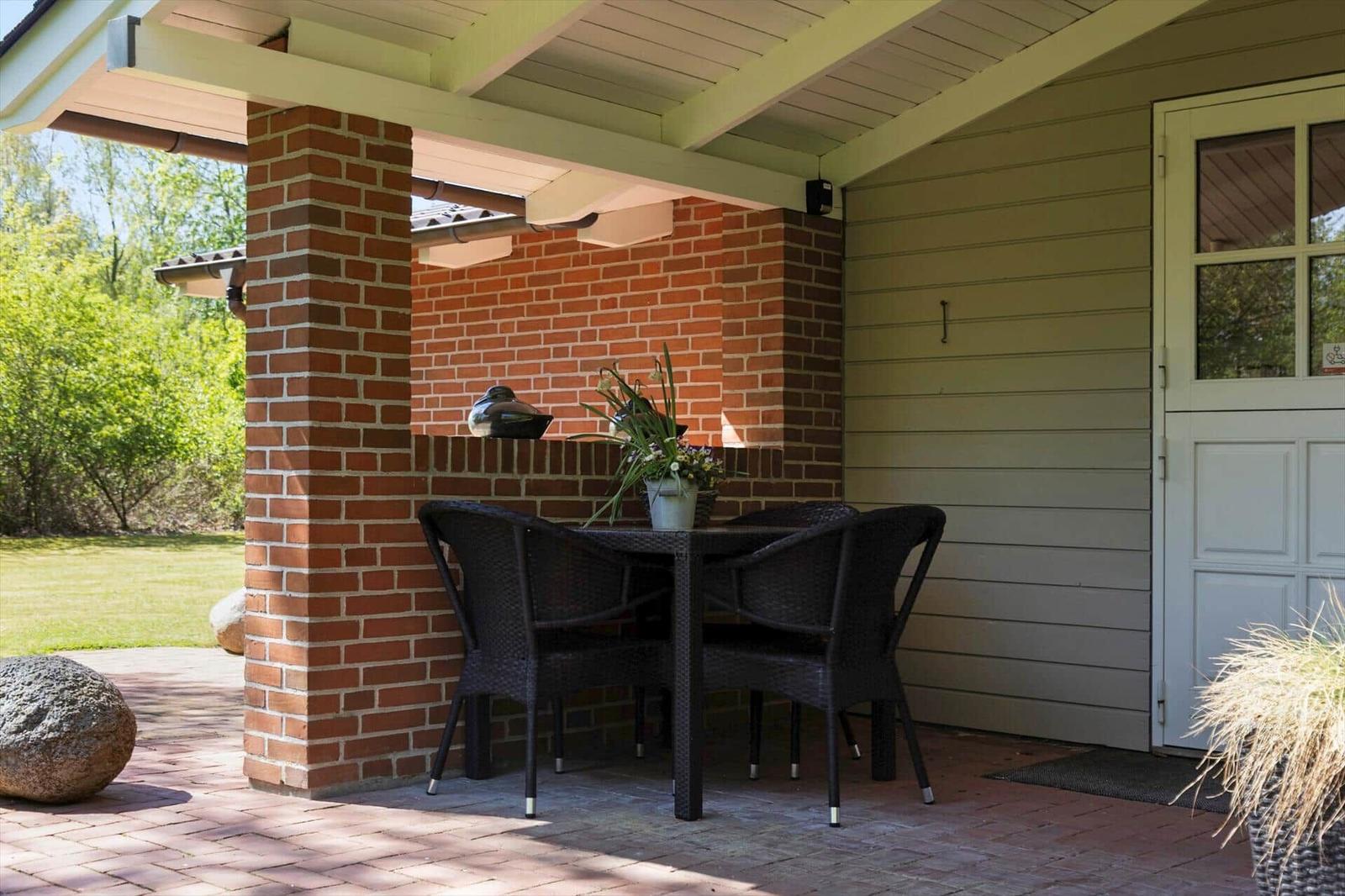 Terrasse mit Tisch und Stühlen, Backsteinwand und Holzdecke. Tür und Garten im Hintergrund.