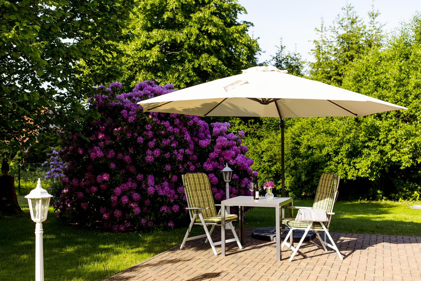 Terrasse mit Tisch, zwei Stühlen und Sonnenschirm. Hintergrund: grüne Bäume und lila Blüten.